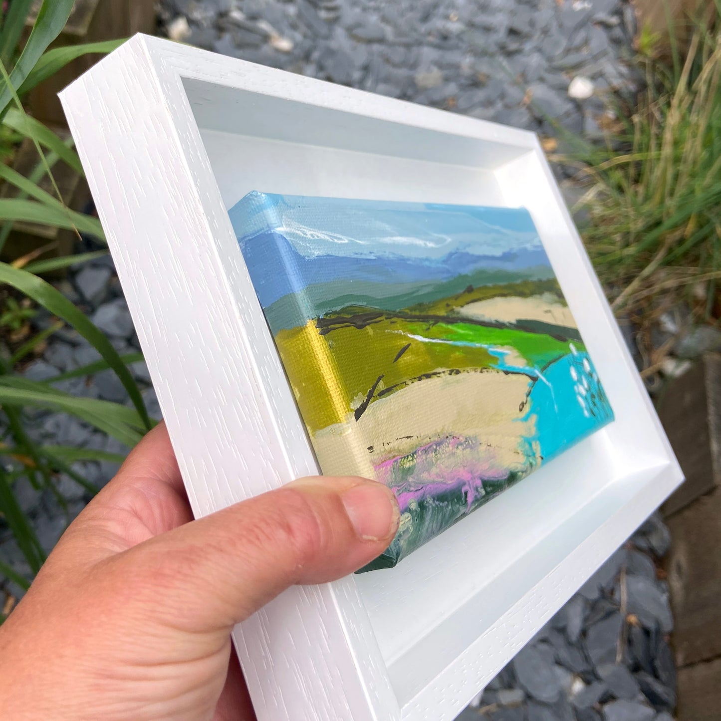 Hand holding a small framed artwork of a landscape with mountains and fields.